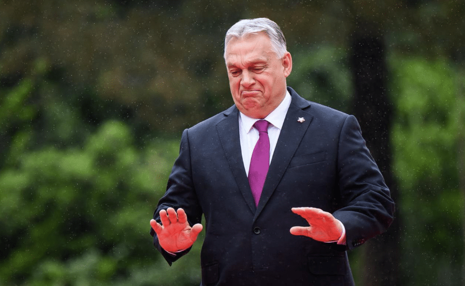 Hungarian Prime Minister Viktor Orbán speaking and pointing during a public address, with EU and German symbols in the background, illustrating his criticism of the European Union and Germany over alleged war preparations.